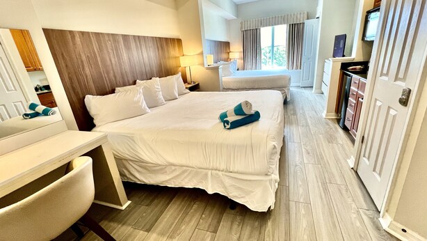 Een hotelkamer met een groot bed, een bureau en een stoel. Er is een raam met gordijnen en een deur aan de rechterkant.