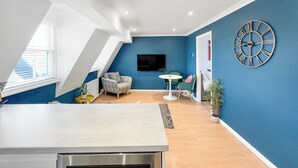 Ein modernes Wohnzimmer mit blauer Akzentwand, einer großen Wanduhr, einem Flachbildfernseher und einem Essbereich mit kleinem Tisch und Stühlen.