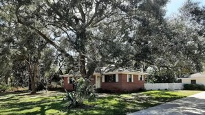 Una casa a un piano con facciata in mattoni, immersa in un prato curato e circondata da alberi di grandi dimensioni.