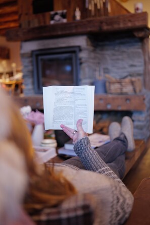 A person reading a book in a cozy room with a stone fireplace and wooden beams.
