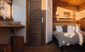 A wooden cabin interior with a bed, bedside lamps, a wooden door, and a window with a view.