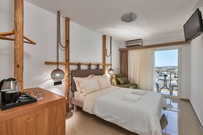 A hotel room with a bed, a wooden side table with a kettle and glasses, a wall-mounted lamp, and a view of the city through the window. - D And D Resort (Hersonissos)