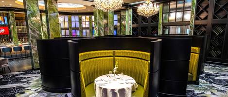 A dining area with a round table, yellow upholstered booths, chandeliers, and a ceiling mural.
