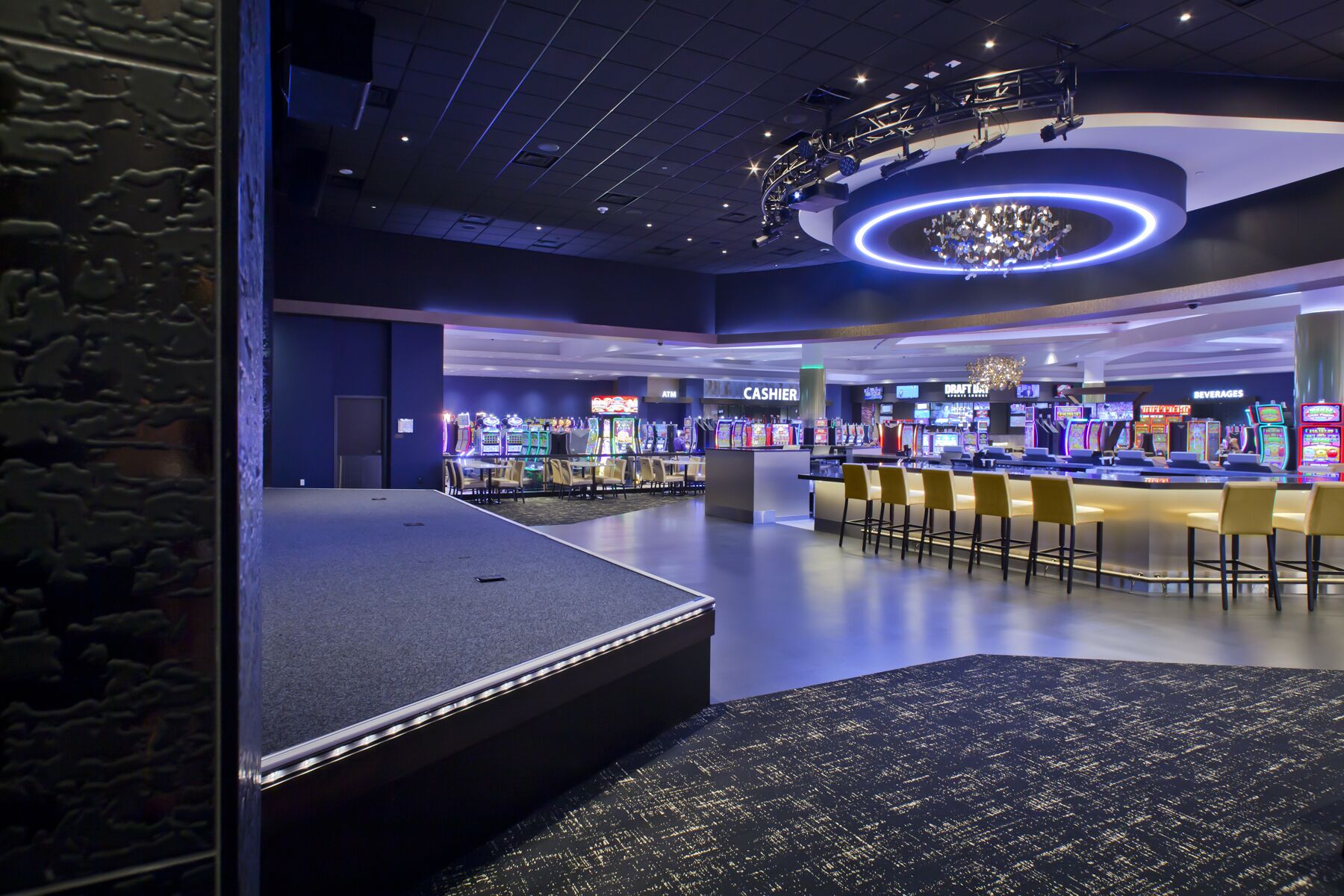 A modern gaming area with multiple arcade machines, a bar area with stools, and a stage for performances.