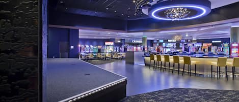 A modern gaming area with multiple arcade machines, a bar area with stools, and a stage for performances.
