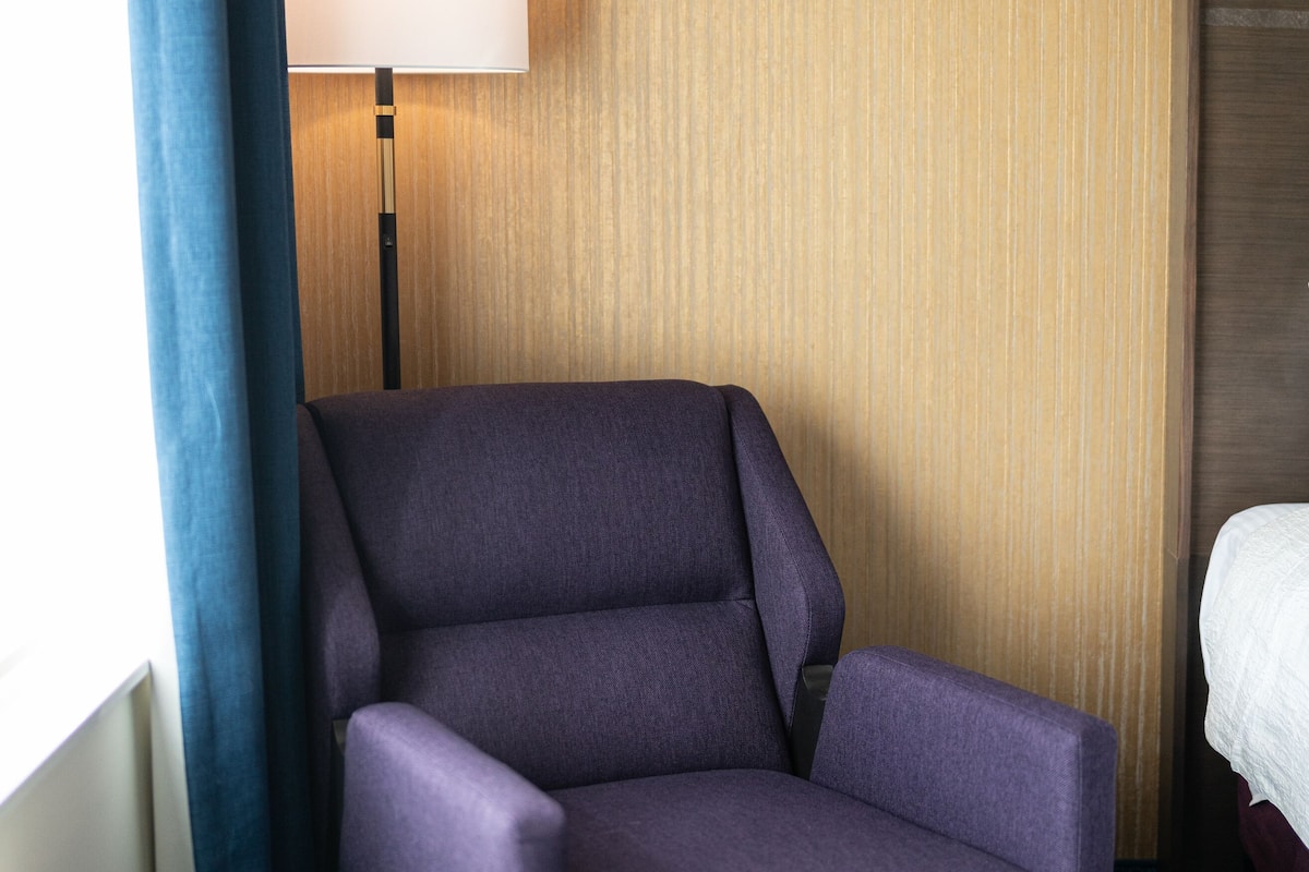A hotel room with a purple armchair, a bedside table, a lamp, and a bed.