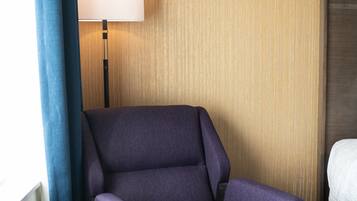 Una habitación de hotel con un sillón morado, una mesita de noche, una lámpara y una cama.