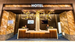 Een receptiebalie in een hotel met een moderne kroonluchter, een bordje met 'SPA' dat binnenkort komt, en twee receptionisten achter hun werkplekken.