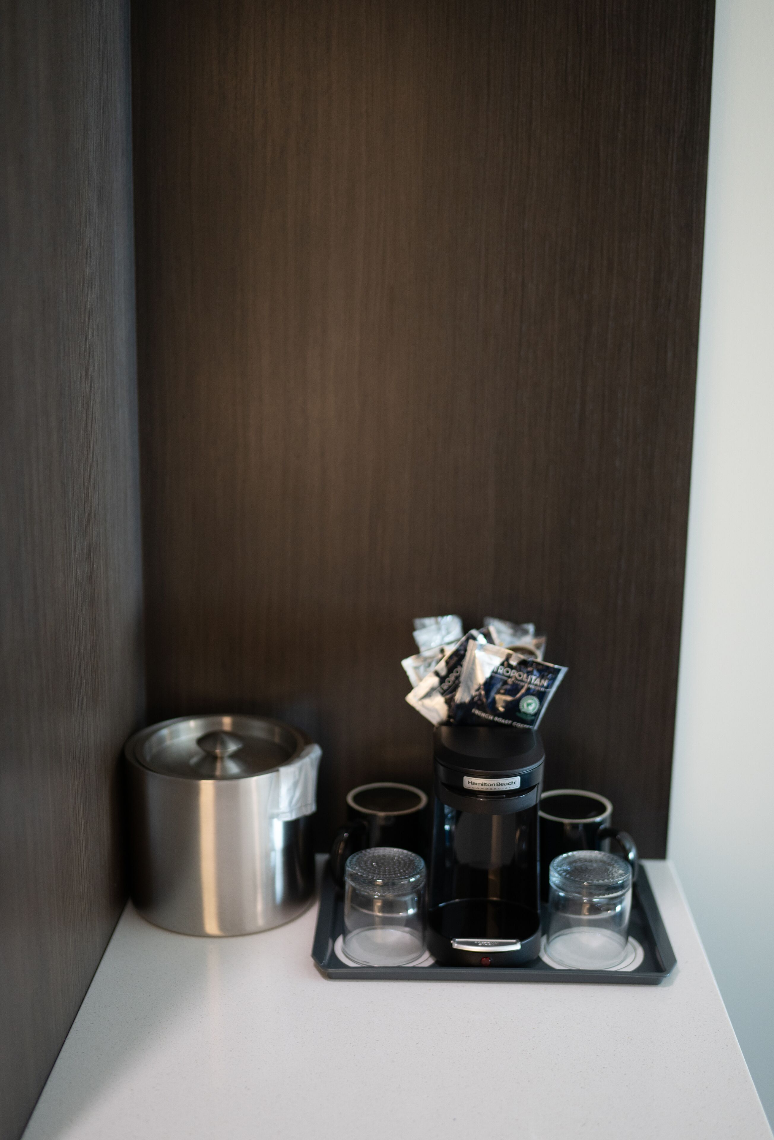 A coffee maker with four cups on a tray, a metal container, and a wooden wall.