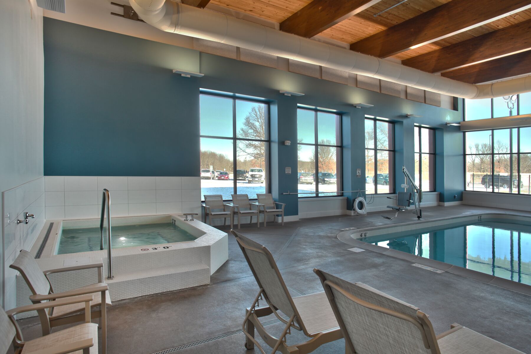 Una piscina interior moderna con jacuzzi, ventanas amplias y vista al estacionamiento.