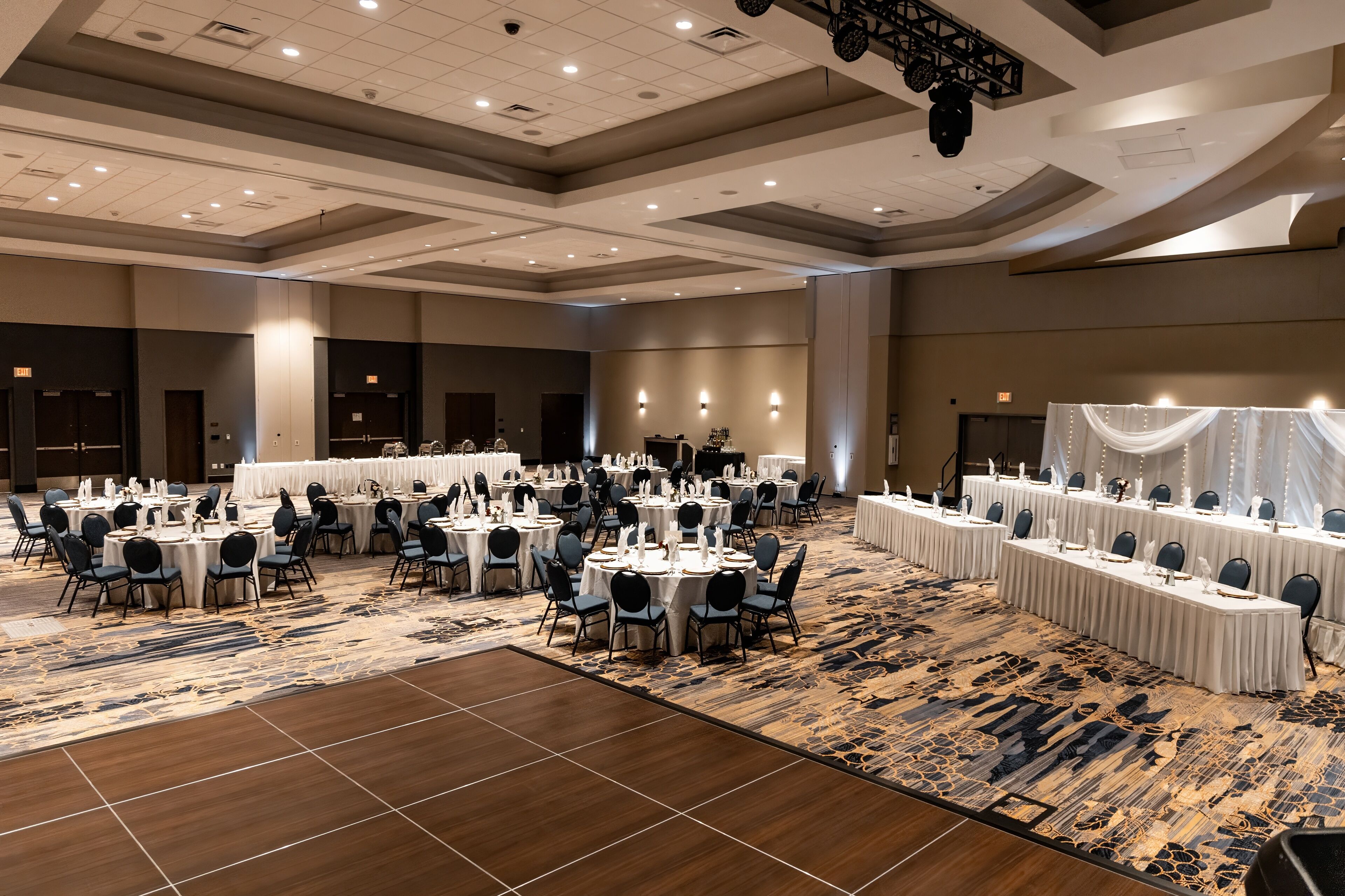 A large banquet hall with round tables and chairs, a patterned carpet, and a high ceiling with recessed lighting.