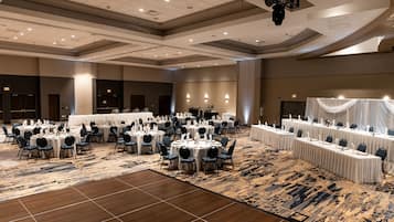 A large banquet hall with round tables and chairs, a patterned carpet, and a high ceiling with recessed lighting.