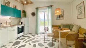 A compact kitchen with teal cabinets, a stove, and a refrigerator. There is a dining area with a round table and chairs, and a sofa with cushions. A hanging light fixture is visible, and a framed quote is on the wall.