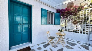 Edifici blanc amb una porta i persianes verda mar, una petita zona d'exterior amb taula i cadires, i una tanca blanca de latici amb plantes florejants.