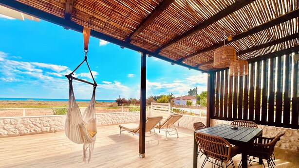 A wooden deck with a table and chairs, a hanging hammock, and a view of the beach.