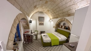 A compact hotel room with a bed, a desk, and a chair. There is a small table with a bottle and glasses, and a wall-mounted shelf with decorative items. - Studio apartment on padreterno street in nardò to relax, 5 minutes from the beach  (Nardò)