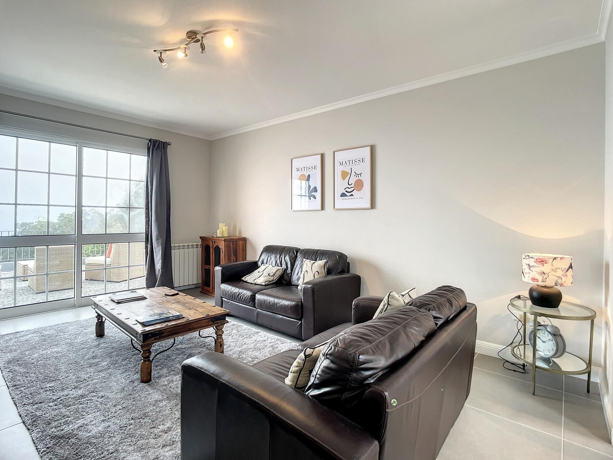 A spacious living room with a large window, a leather sofa, a coffee table, and two framed artworks on the wall.