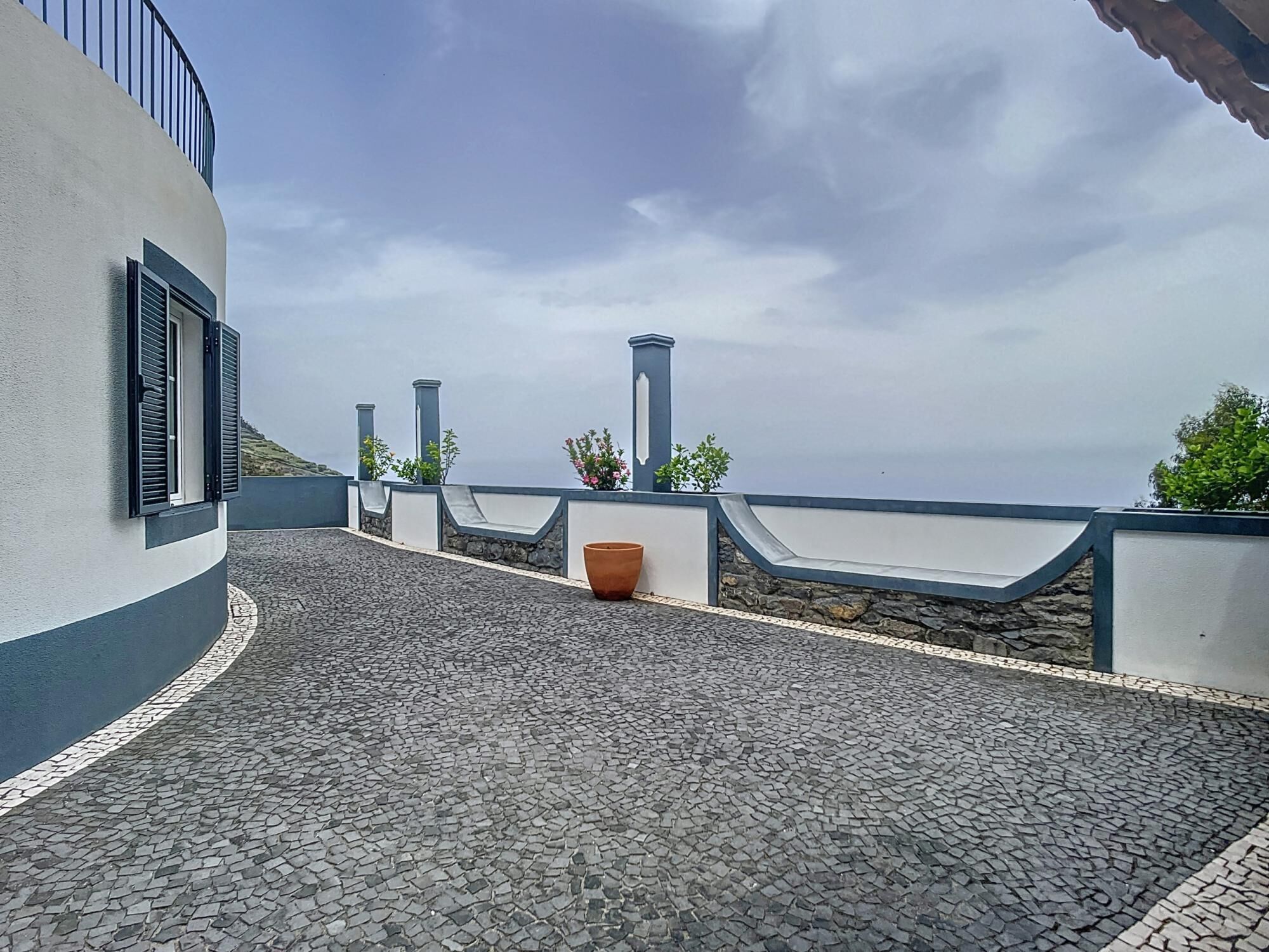 A paved terrace with a stone floor, a white building with black shutters, and a view of the sea under a cloudy sky.