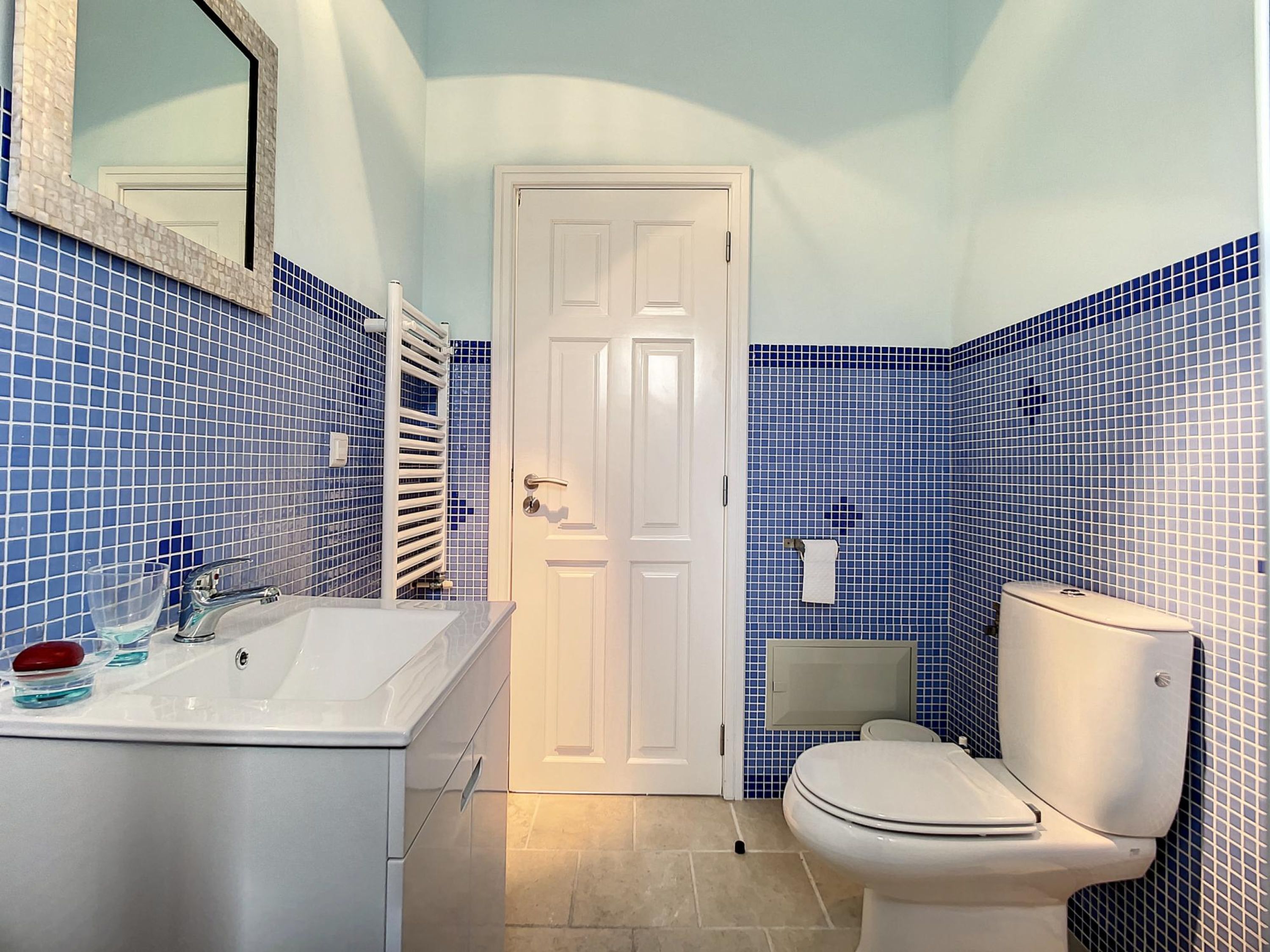 Un baño con un lavamanos blanco, un espejo, un inodoro blanco y paredes con azulejos azules.