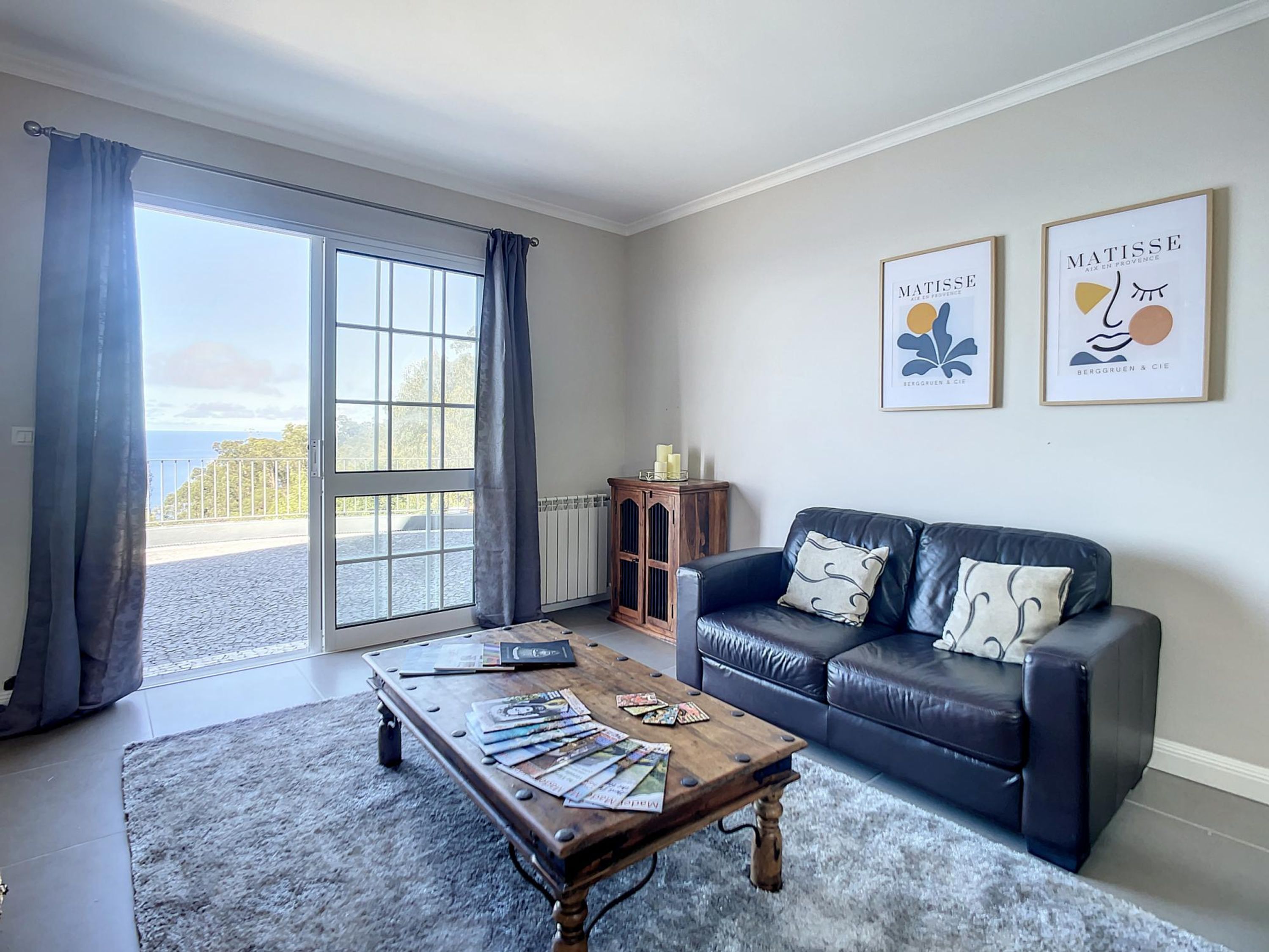 Un salon avec un canapé en cuir noir, une table basse en bois sur laquelle sont posés des magazines, un radiateur et une vue sur la mer par une grande fenêtre.