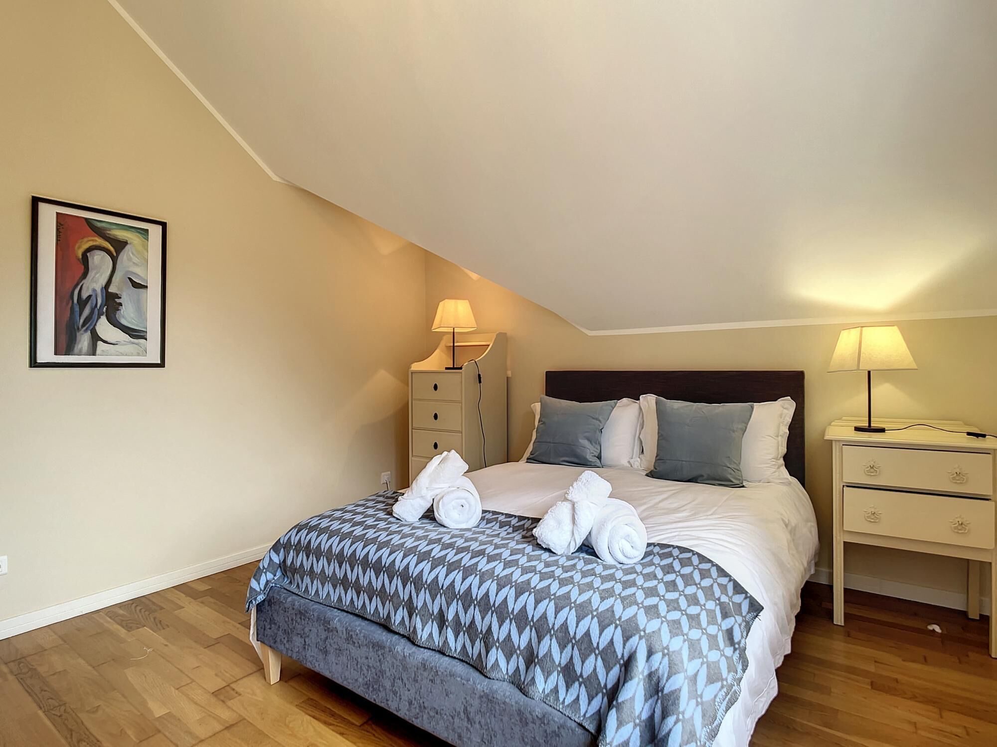 A bedroom with a bed, two bedside tables, a nightstand, a painting, and a wooden floor.