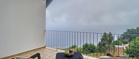 A balcony with wicker furniture and a table, overlooking a body of water and greenery.