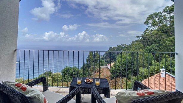 Un balcón con mobiliario de mimbre, una mesa con un vaso de jugo de naranja y vistas al mar y la vegetación.