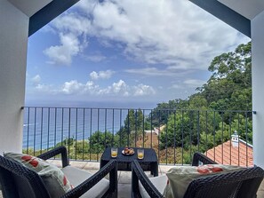 A balcony with wicker furniture, a table with a glass of orange juice, and a view of the sea and greenery. - Vinhaticos House by Atlantic Holiday (Porto Moniz)