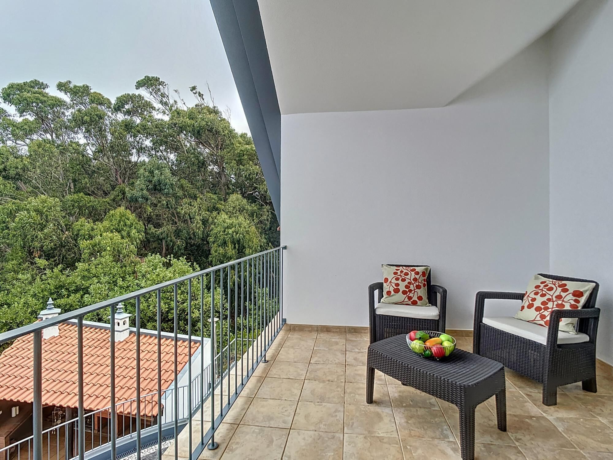 A balcony with wicker furniture, a tiled floor, and a view of a red-roofed building and green trees.