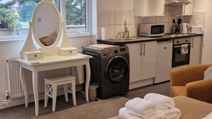 A compact, well-lit room with a washing machine, a microwave, and a sink. There's a small table with a mirror and a chair, and a sofa with towels. - Charming Studio Rutland Point Holiday or Work (Oakham)