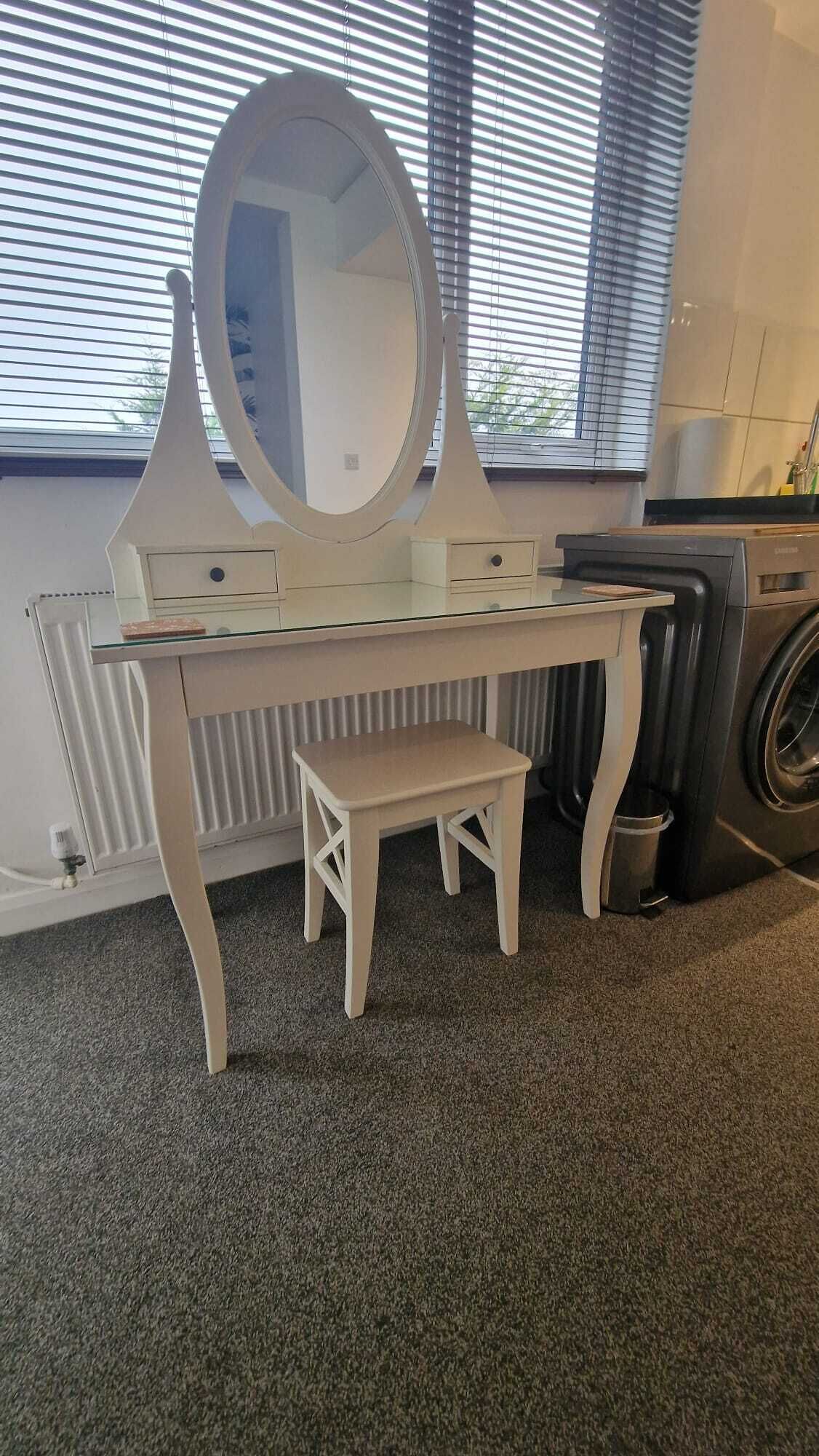A white vanity with a large mirror and a small stool, positioned in front of a radiator and a washing machine.