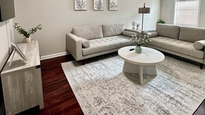 Un salon moderne avec un canapé beige, une table basse ronde et un tapis blanc. Trois œuvres d’art florales encadrées ornent le mur, ainsi qu’une lampe sur pied et une petite plante en pot.