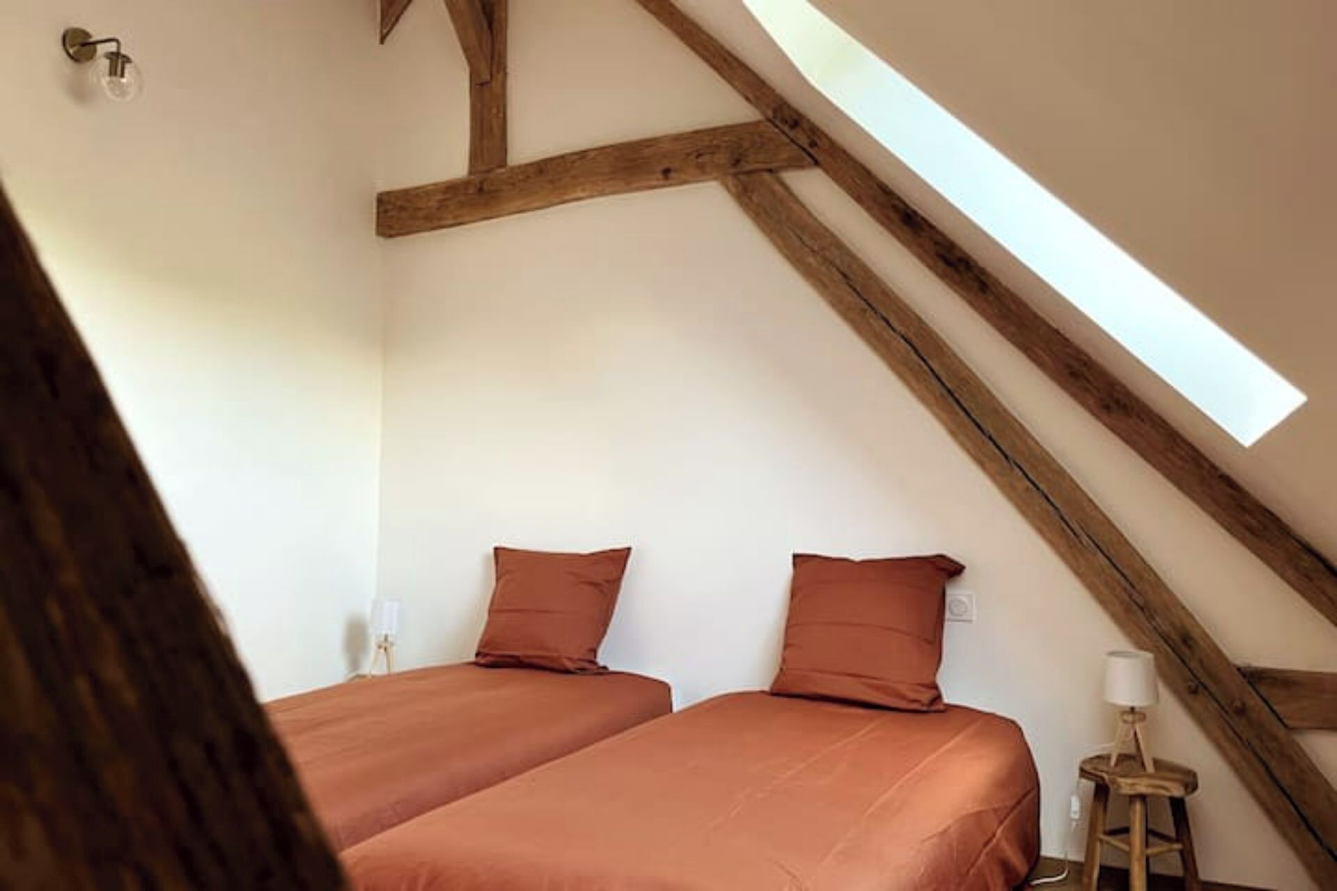 Une chambre avec deux lits, des poutres apparentes, une lucarne et une petite table avec une lampe.