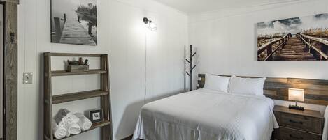 Une chambre à coucher bien rangée, avec un lit, une table de chevet dotée d’une lampe, une étagère en bois avec des serviettes et deux tableaux accrochés au mur.