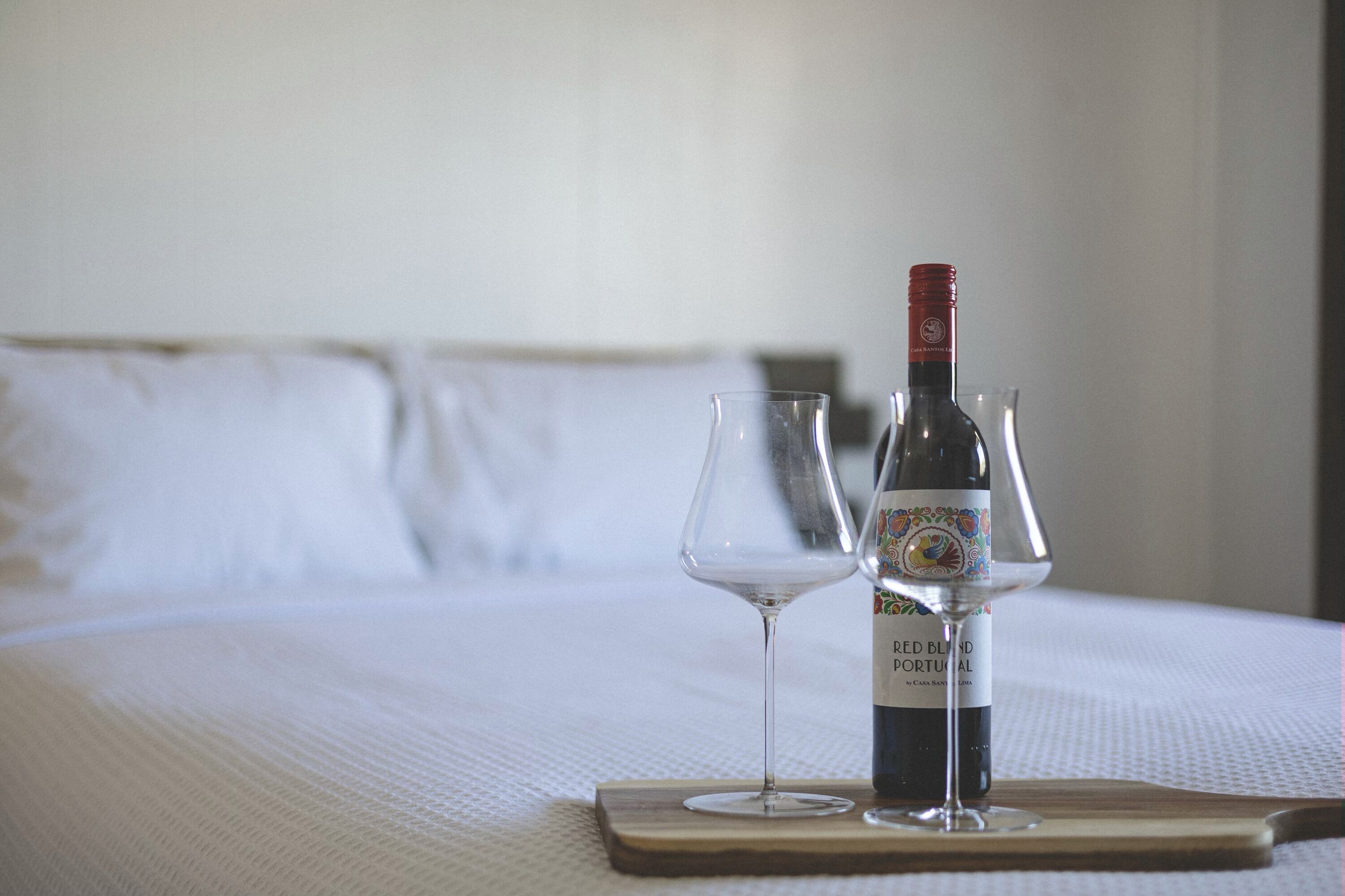 A bed with a wine bottle and two glasses on a wooden tray.