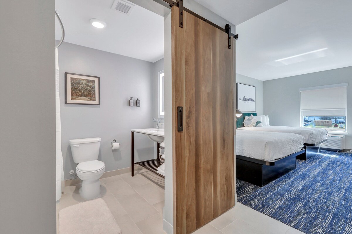 A modern hotel room with a wooden sliding door, a white toilet, a sink, a bed, and a blue carpet.