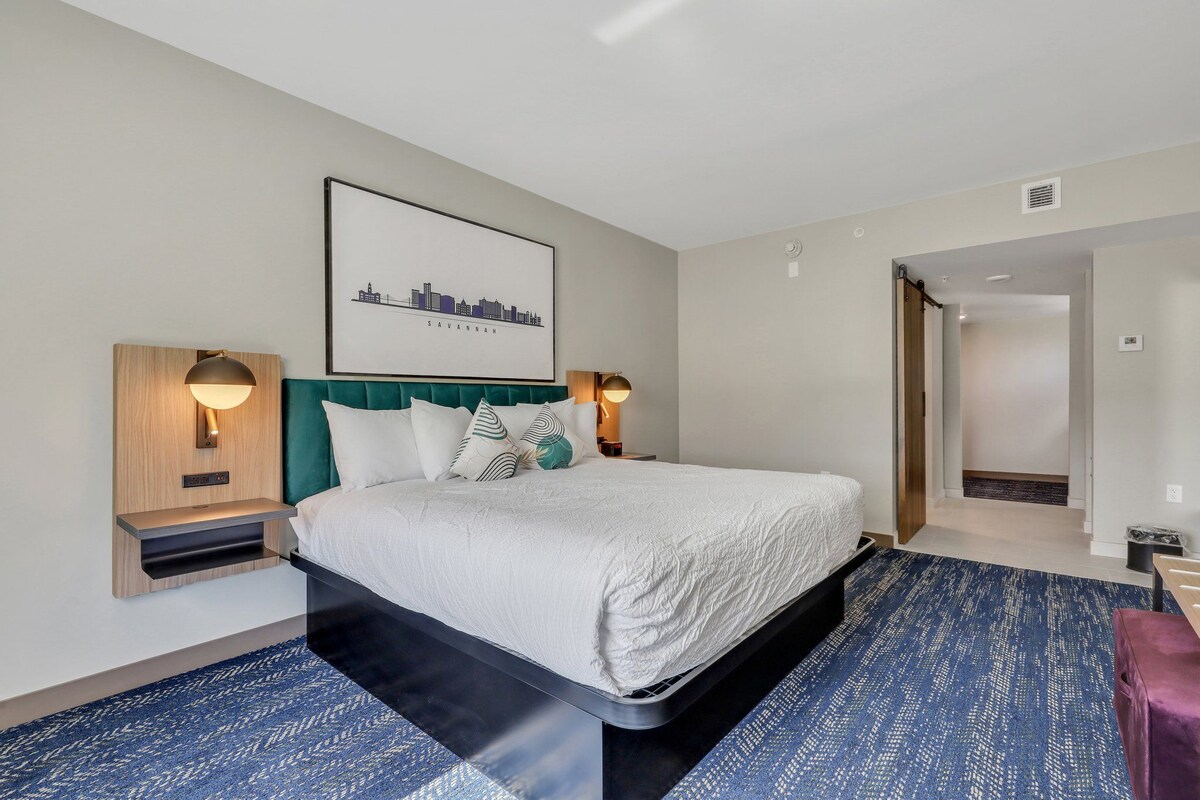 A hotel room with a bed, two bedside tables, a wall-mounted light, a framed cityscape picture, and a blue patterned carpet.