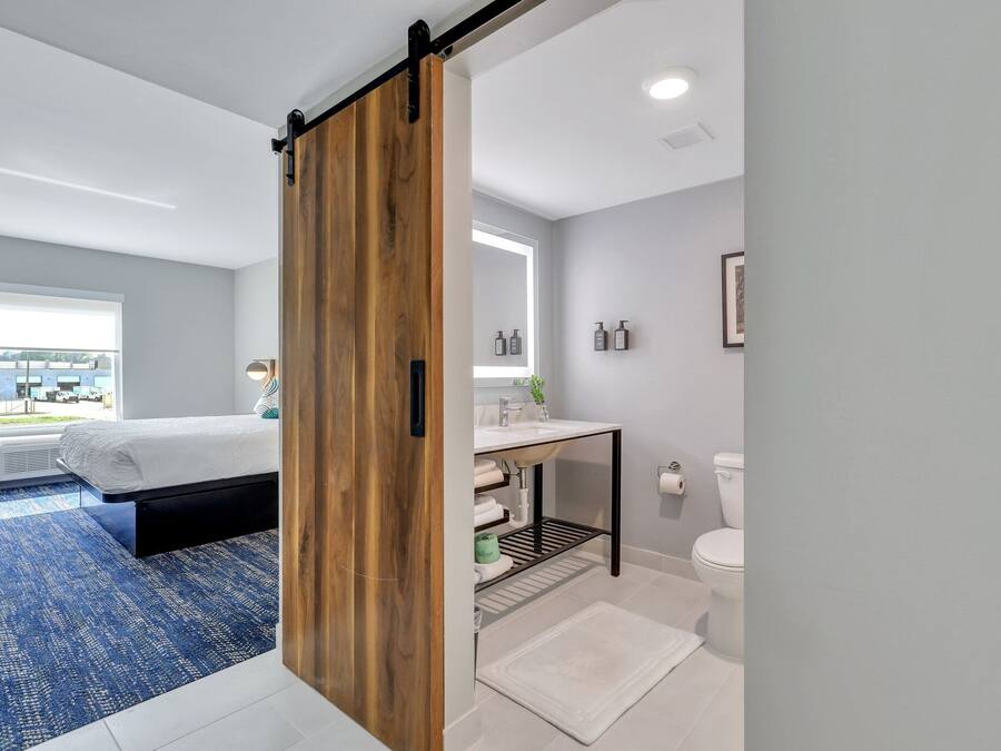 A modern bedroom with a large bed, a sliding wooden door, a bathroom with a sink and toilet, and a view of the outdoors.