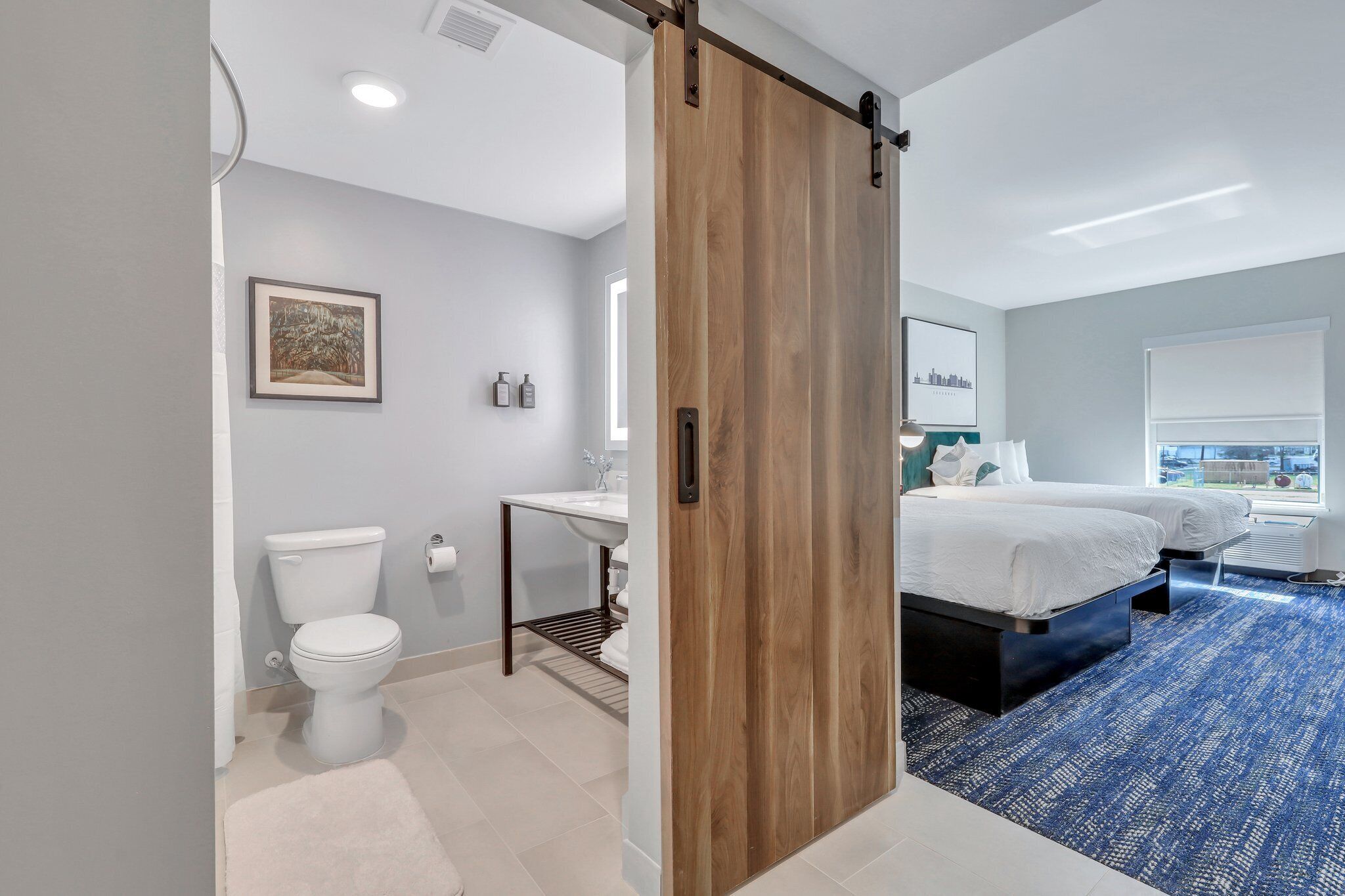 Une chambre d’hôtel moderne avec une porte coulissante en bois, des toilettes blanches, un lavabo, un lit et une moquette bleue.