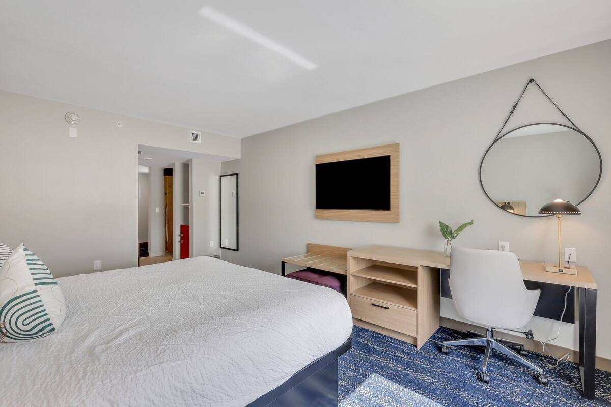 A hotel room with a large bed, a desk with a chair, a mirror, and a small plant.