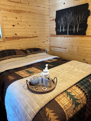 A wooden cabin room with a bed, a tray with toiletries, and a wall art of trees.