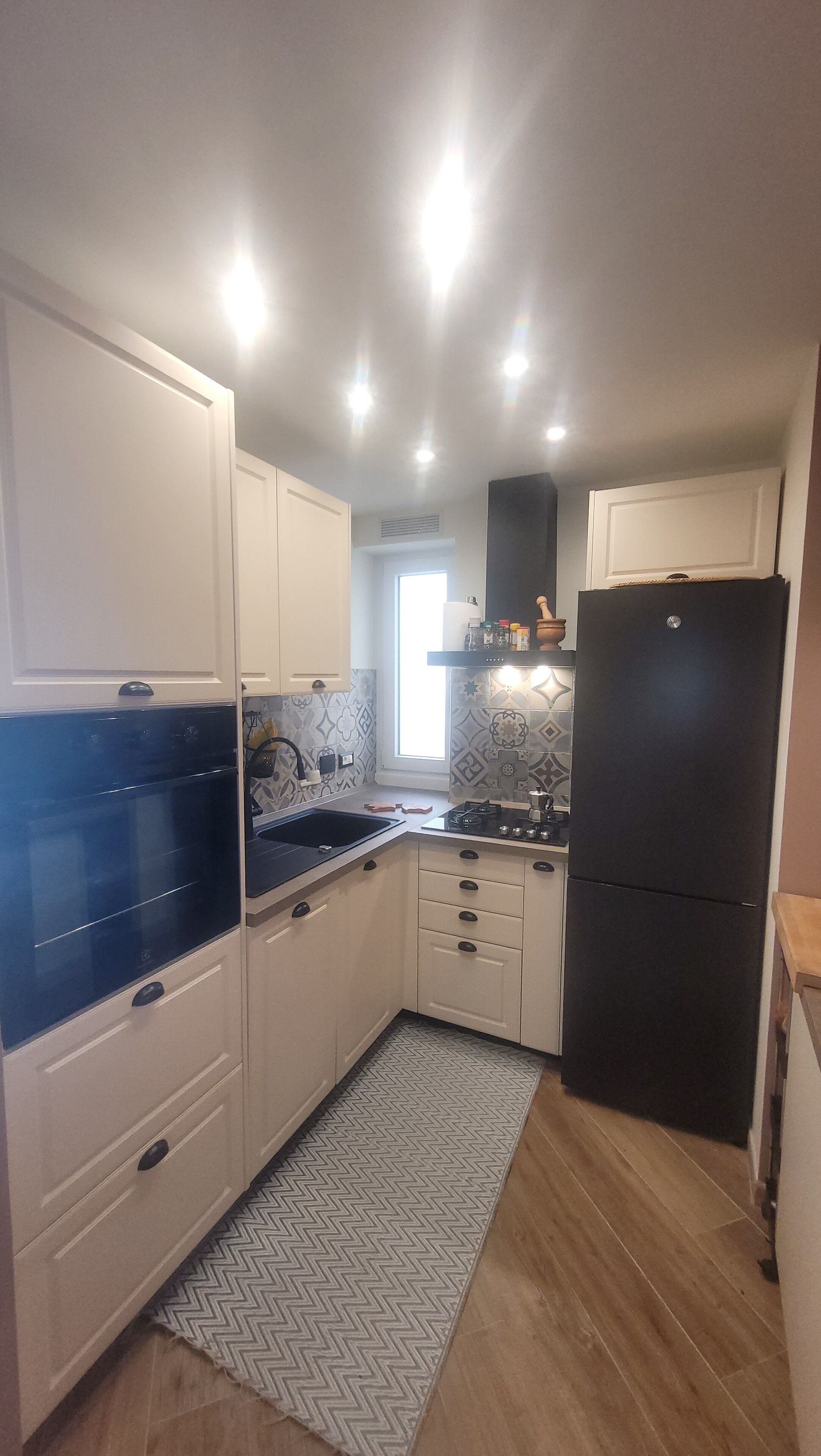 Cocina compacta con muebles blancos, un refrigerador negro y un salpicadero estampado.
