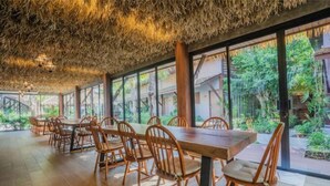 A spacious dining area with wooden tables and chairs, large windows offering views of greenery, and a thatched ceiling.