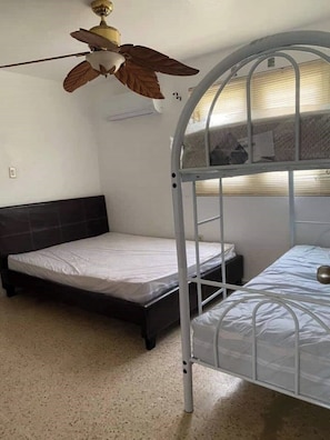 Une chambre avec un lit superposé, un lit simple, un ventilateur de plafond et une fenêtre avec des stores.