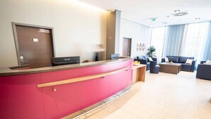 A hotel reception area with a pink counter, a reception desk, a seating area with sofas and a coffee table, and a large window with curtains. - Hotel Orangerie (Vienna)