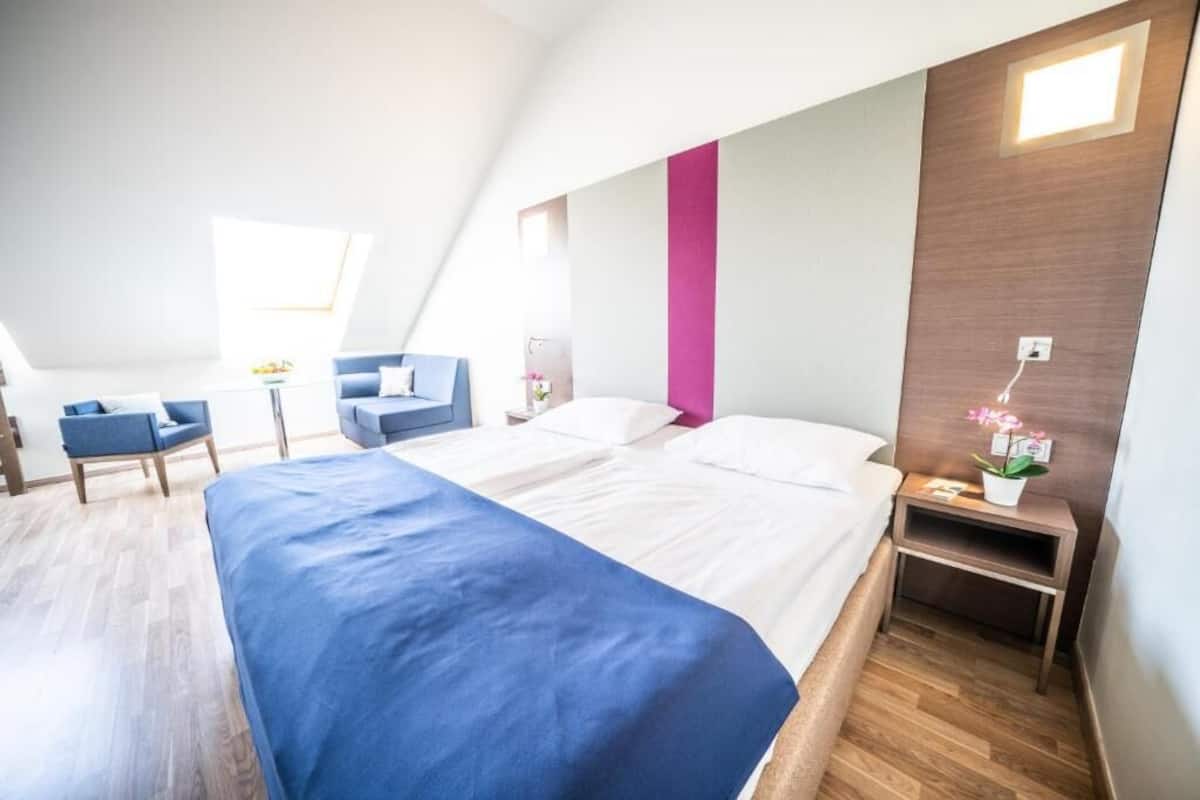 A modern hotel room with a large bed, a desk, a chair, and a small table with a plant.