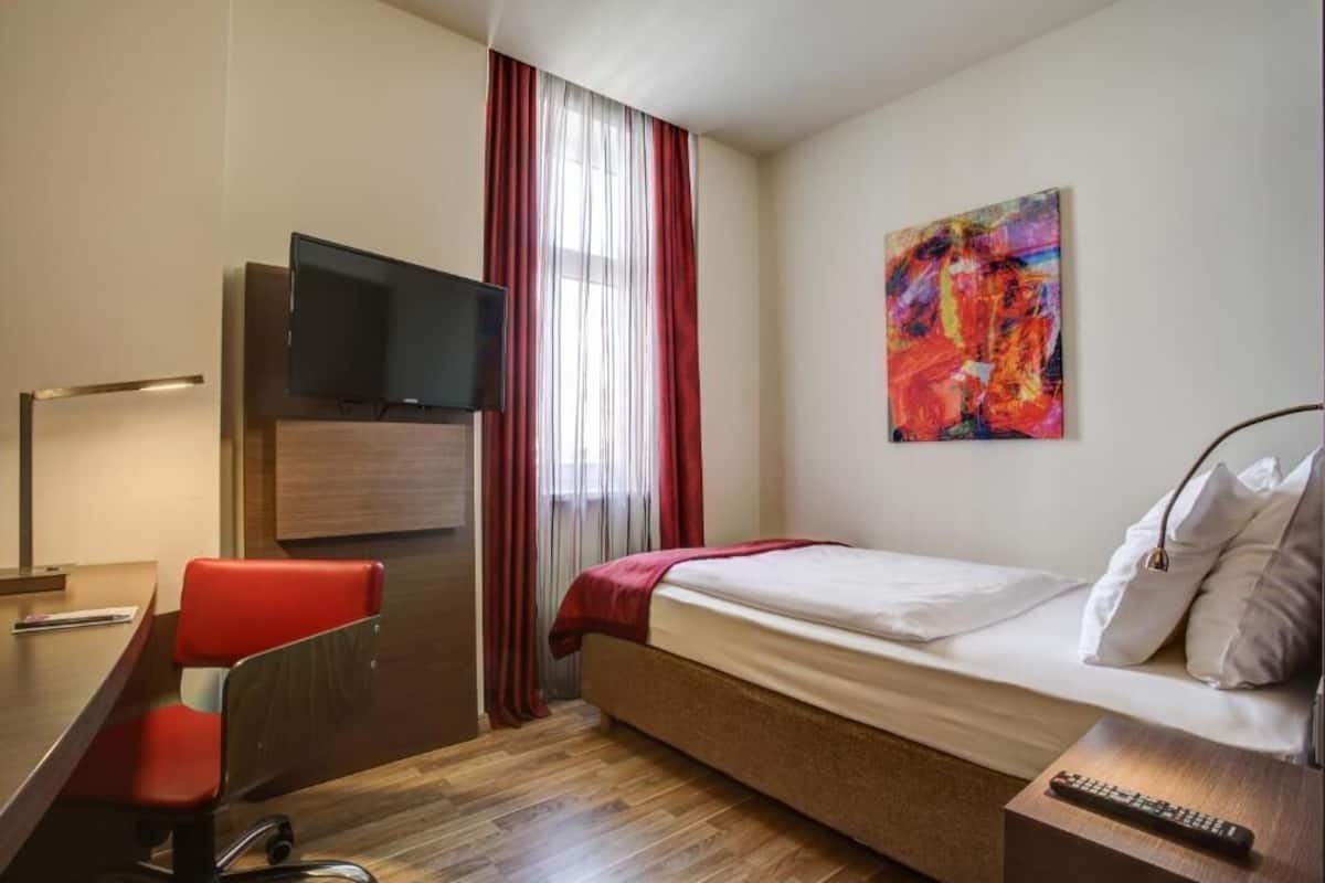 A hotel room with a bed, a desk with a chair, a TV, a painting on the wall, and a window with curtains.