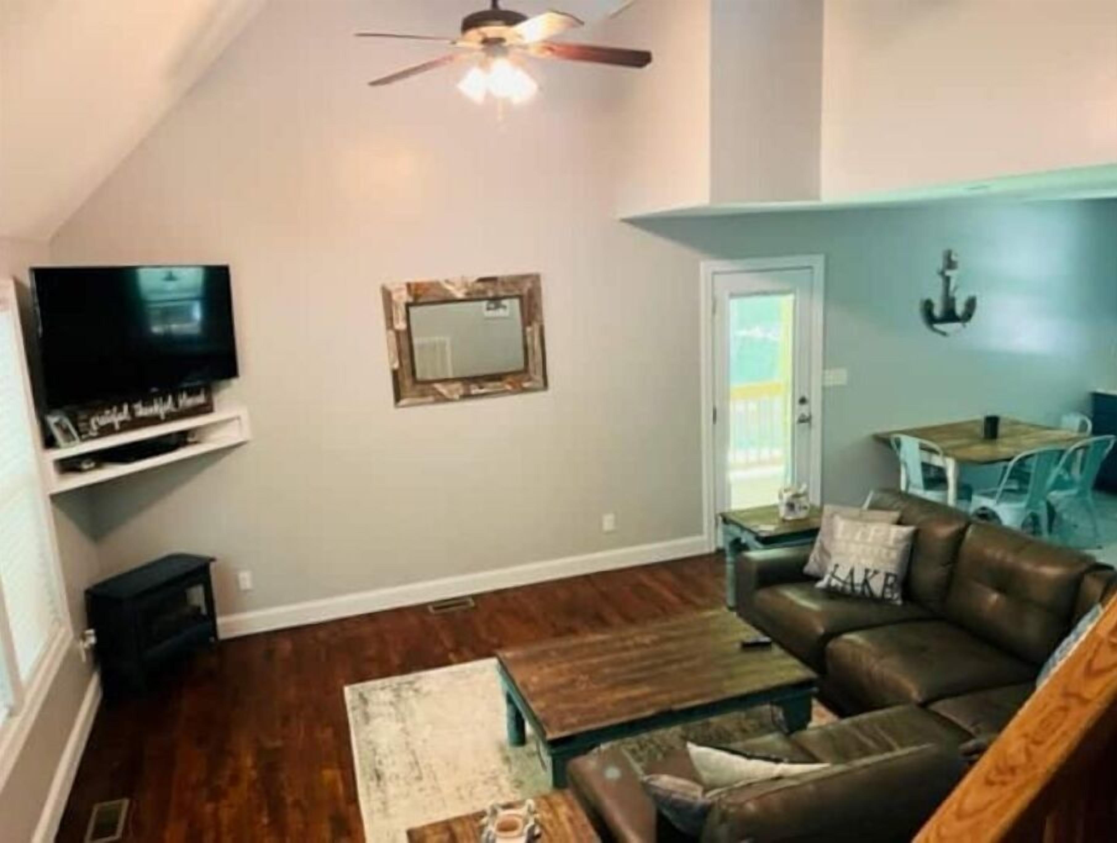 A living room with a brown leather sofa, a coffee table, a television on a stand, a ceiling fan, and a small table with chairs.
