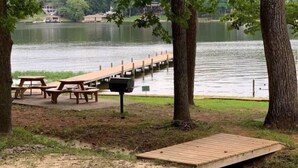 Un área de picnic junto al lago con bancos de madera y una parrilla, rodeada de árboles y un muelle que se adentra en el agua.