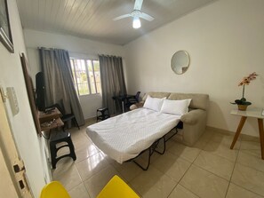 Habitación de hotel pequeña con cama, escritorio, silla, mesita con planta en maceta y ventilador de techo.
